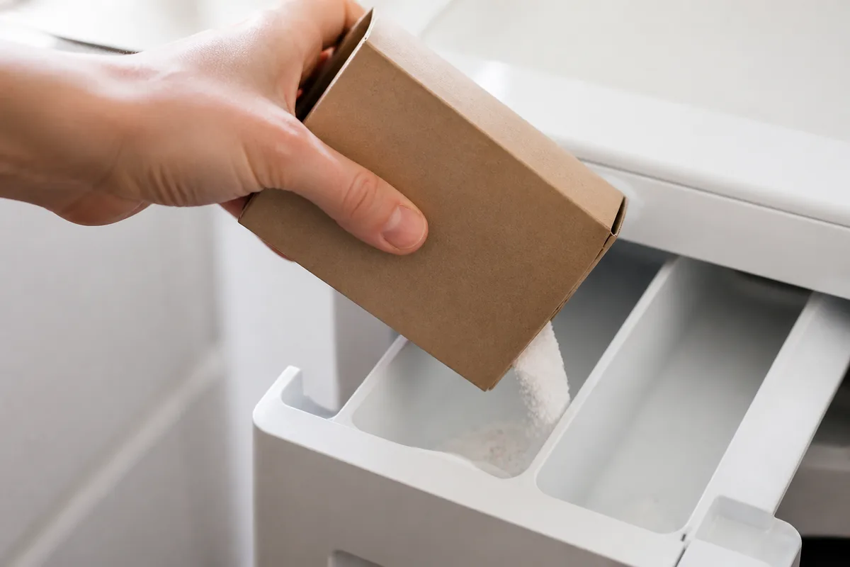 Main versant une poudre blanche granuleuse d&rsquo;une boîte en carton dans le bac d&rsquo;un lave-linge